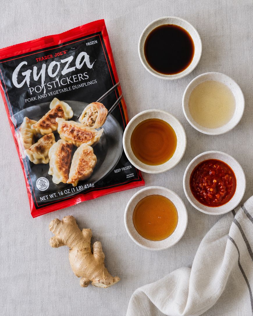 A package of Trader Joe’s Gyoza Potstickers is surrounded by five small bowls of various dipping sauces, a piece of ginger root, and a striped napkin on a light, textured surface.