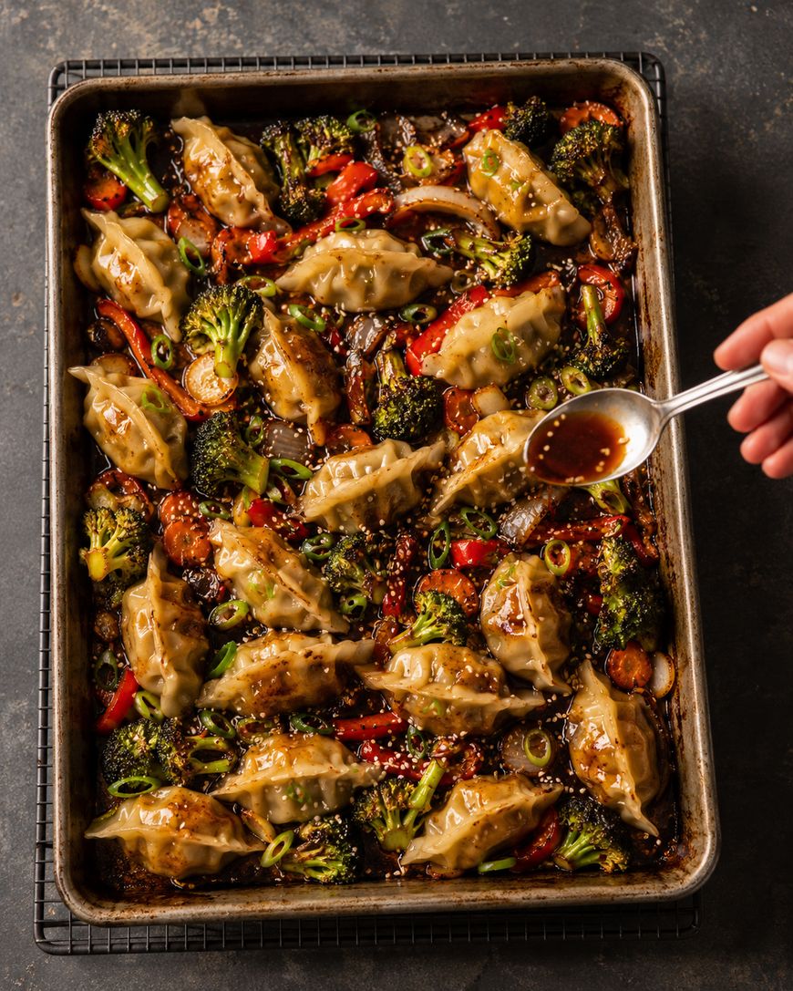 A hand drizzles sauce over a sheet pan filled with cooked dumplings, broccoli, red bell peppers, and green onions, all arranged on a dark baking tray.