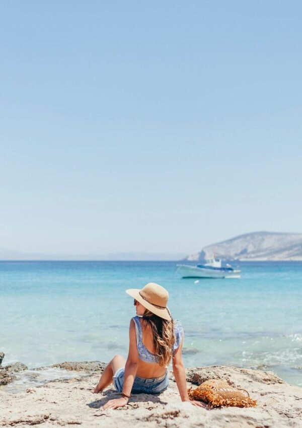 A woman wearing a sunhat and summer clothes sits on a rocky shore, facing the turquoise sea. A boat floats in the distance, and mountains are visible under a clear, blue sky.