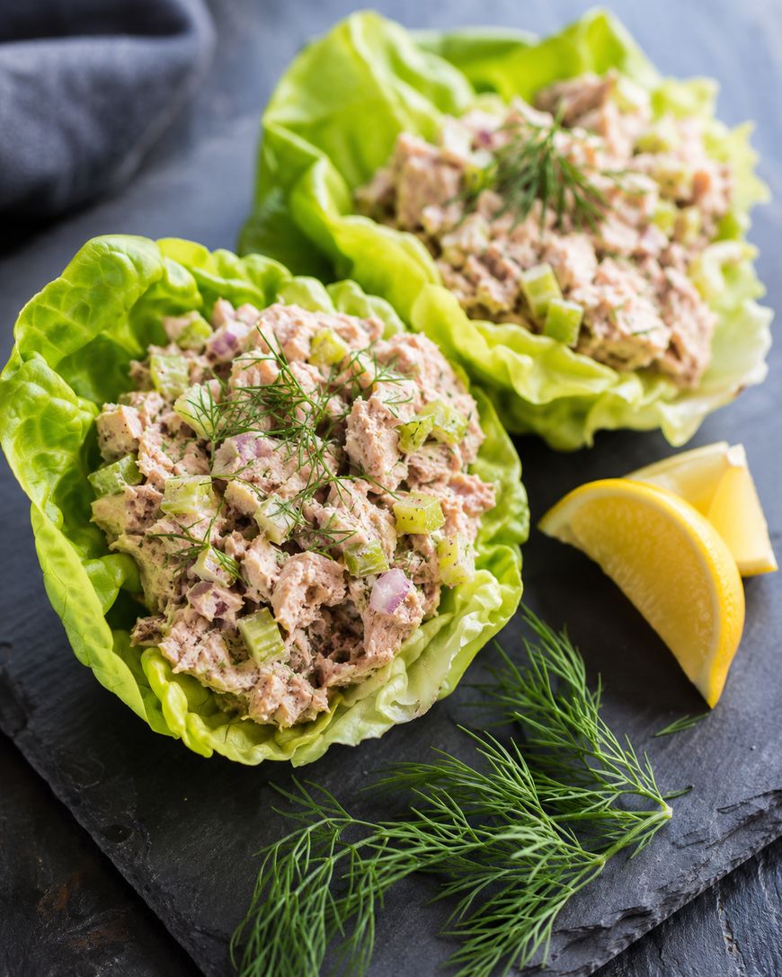 Two lettuce leaves filled with tuna salad, diced celery, and red onion, garnished with fresh dill, served on a dark slate with lemon wedges and dill sprigs on the side.