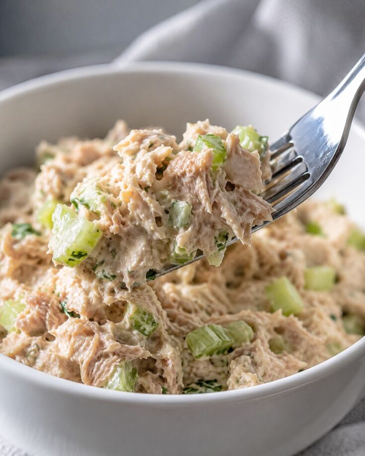 A close-up of a bowl of tuna salad mixed with diced celery. A fork is lifting a portion of the creamy salad from the bowl, highlighting the texture and chunks of celery in the mixture.
