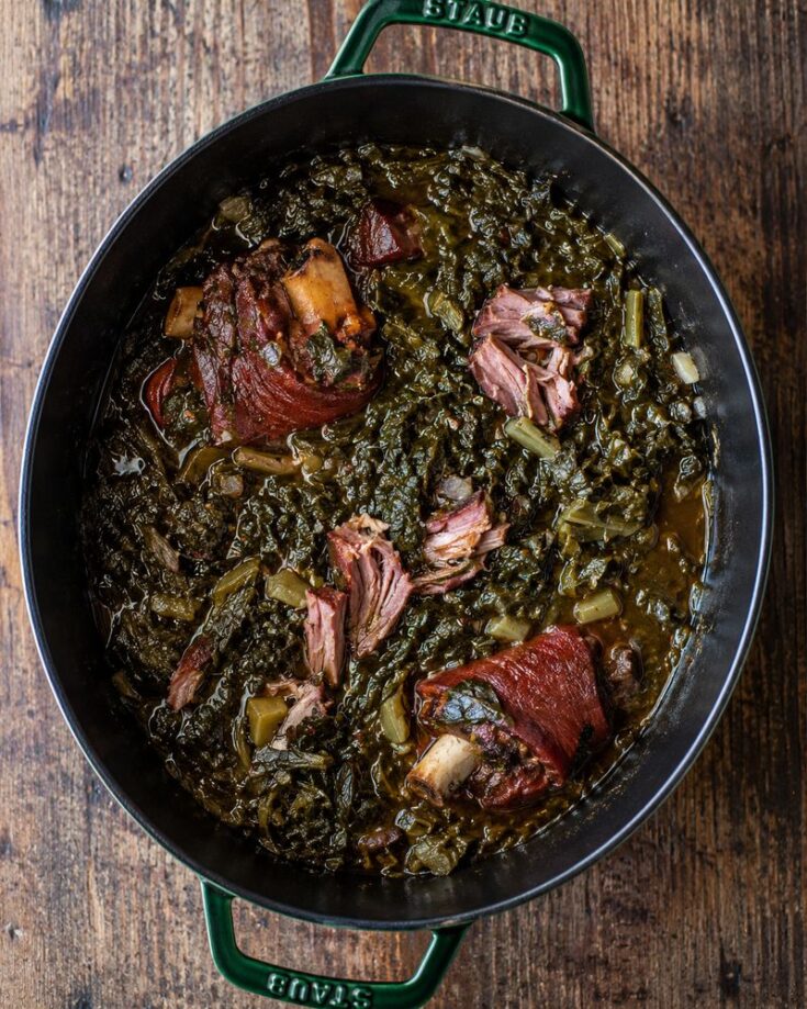 A pot of cooked collard greens mixed with chunks of smoked meat, likely ham hocks, in a savory broth, sits on a wooden table—perfect inspiration for your next delicious turnip greens recipe.