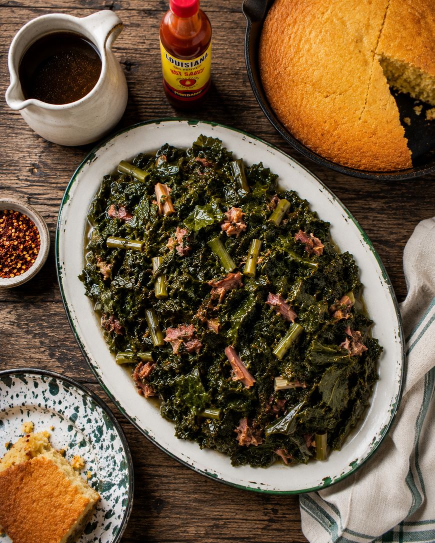A platter of cooked collard greens with ham sits on a rustic table beside a dish of cornbread, a plate with a cornbread slice, a bottle of hot sauce, a small bowl of chili flakes, and a jug of brown sauce.