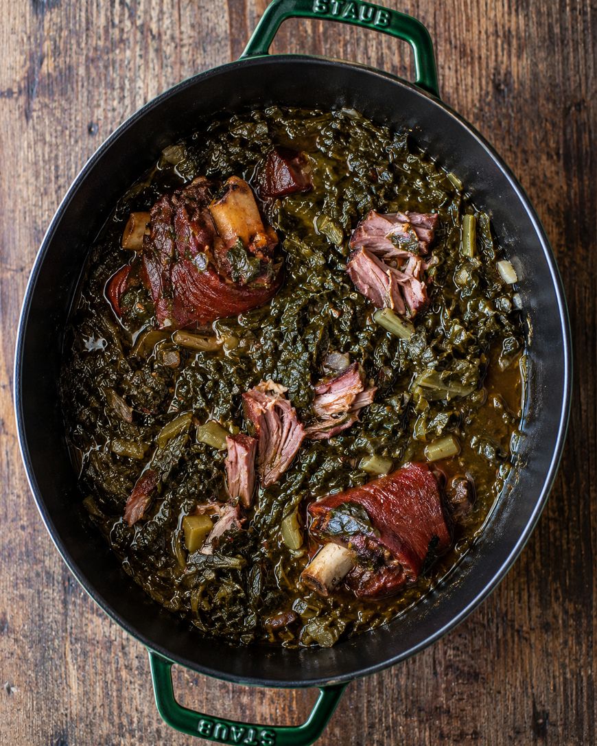 A black pot filled with cooked collard greens and smoked ham hocks sits on a wooden surface. The greens are tender and dark, with pieces of ham hock visible throughout the dish.