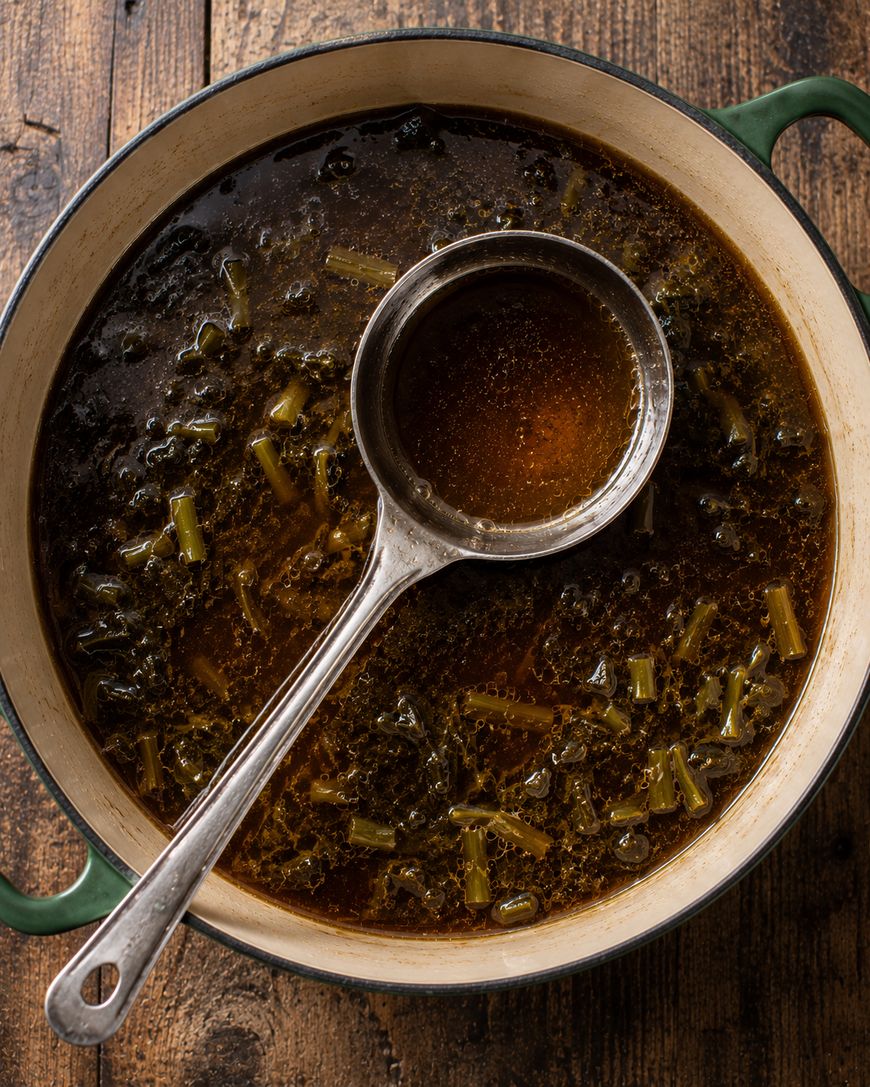 A large pot of dark vegetable broth with chopped greens and a ladle resting inside, sitting on a rustic wooden table.