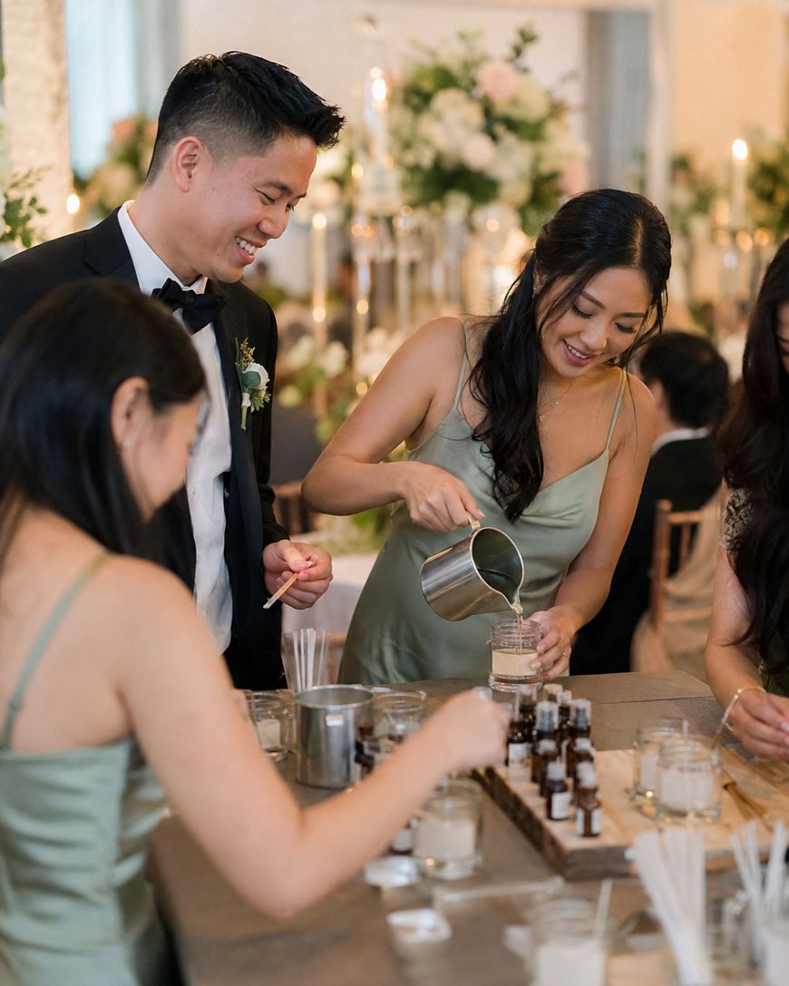 Three people in formal attire smile and interact at a table, where one woman pours liquid from a metal pitcher into a glass jar. The table holds small bottles and candle-making supplies. Elegant decor is seen in the background.