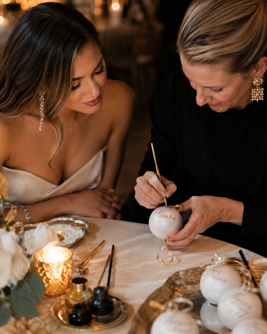 Two women sit at a table decorated with candles and flowers. One woman paints the name Laura on a white ornament while the other woman watches closely, both appearing focused and engaged in the festive activity.