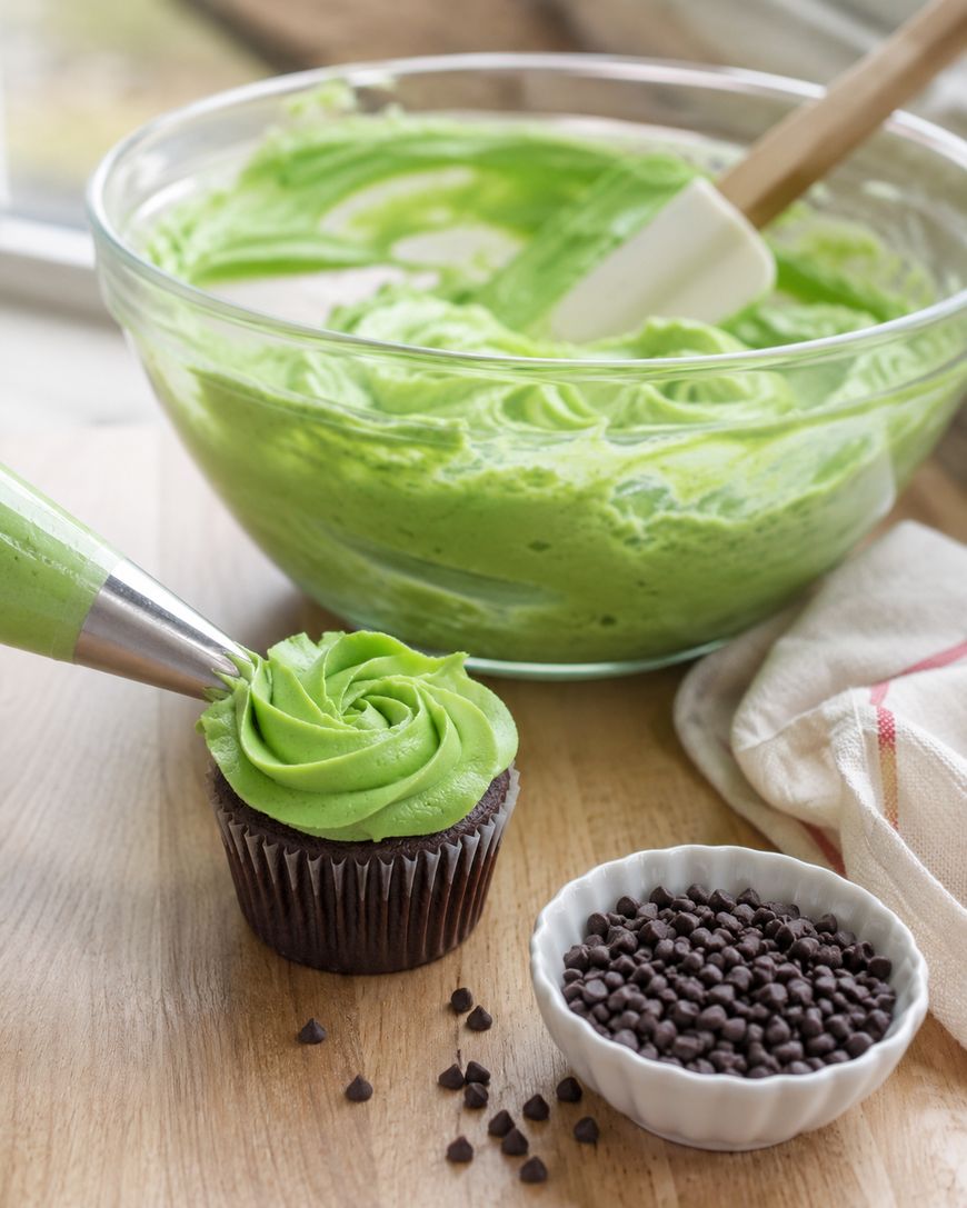A chocolate cupcake is being decorated with green frosting from a piping bag. Behind it, a glass bowl with green frosting and a spatula sits on the table, next to a small bowl of mini chocolate chips.