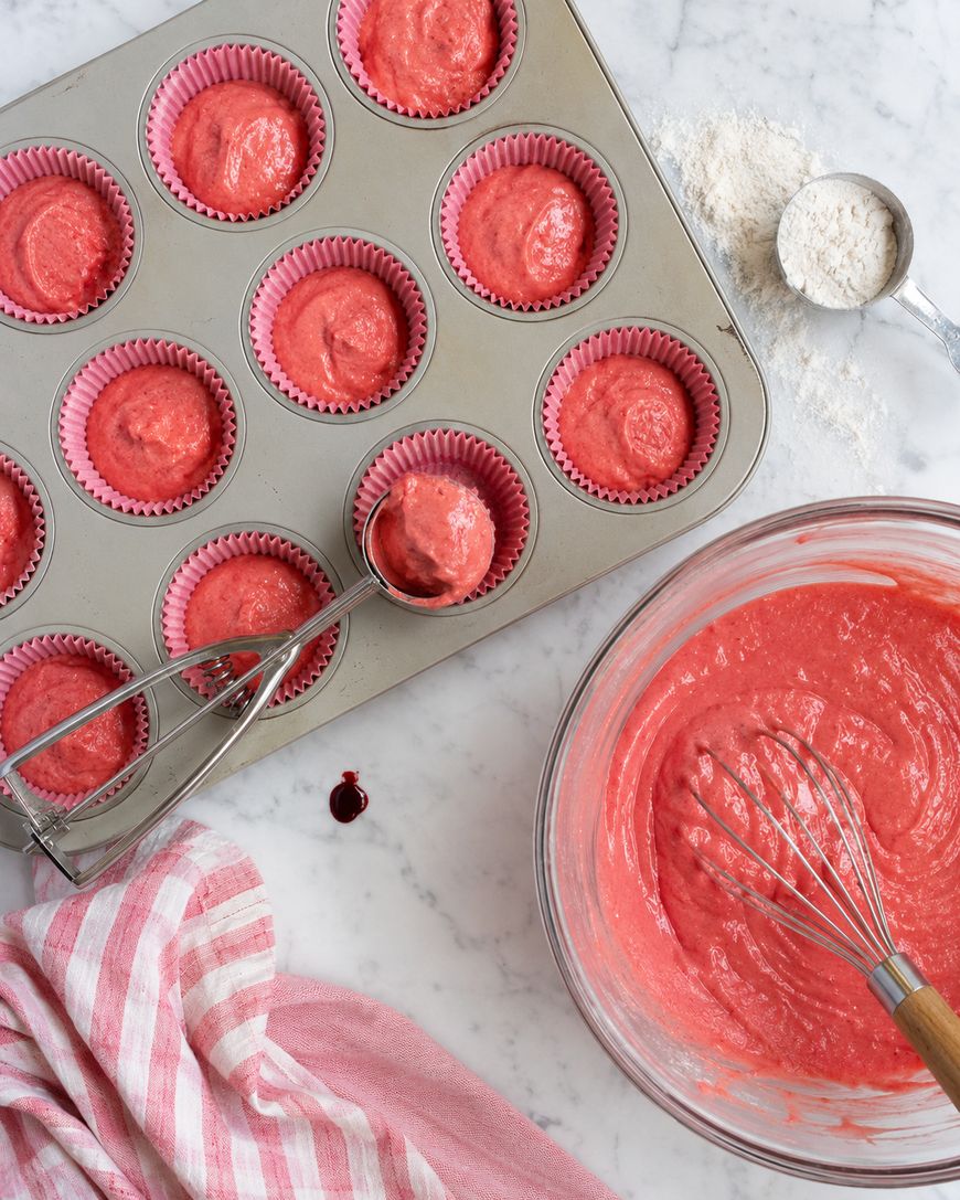 A muffin tin lined with pink paper cups is filled with pink cupcake batter. A metal scoop rests on the tin. Nearby, a glass bowl holds more batter with a whisk, and a striped towel and spoonful of flour are on the marble counter.