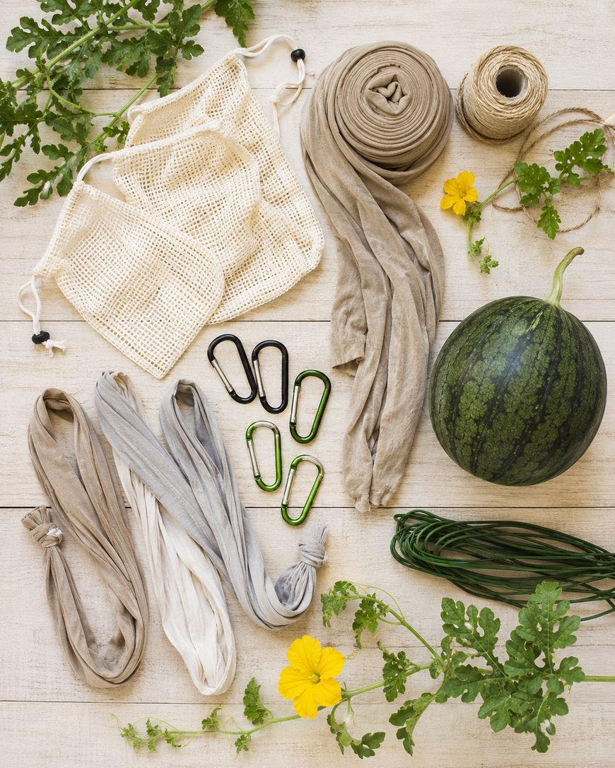 Reusable mesh bags, cloth strips, twine, carabiners, green twist ties, a small watermelon, yellow flowers, and green vines are arranged on a light wooden surface.