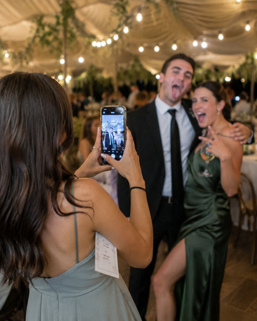 A woman takes a photo with her phone of a smiling man in a suit and a woman in a green dress posing playfully at a lively, elegant event under a tent decorated with string lights and greenery.