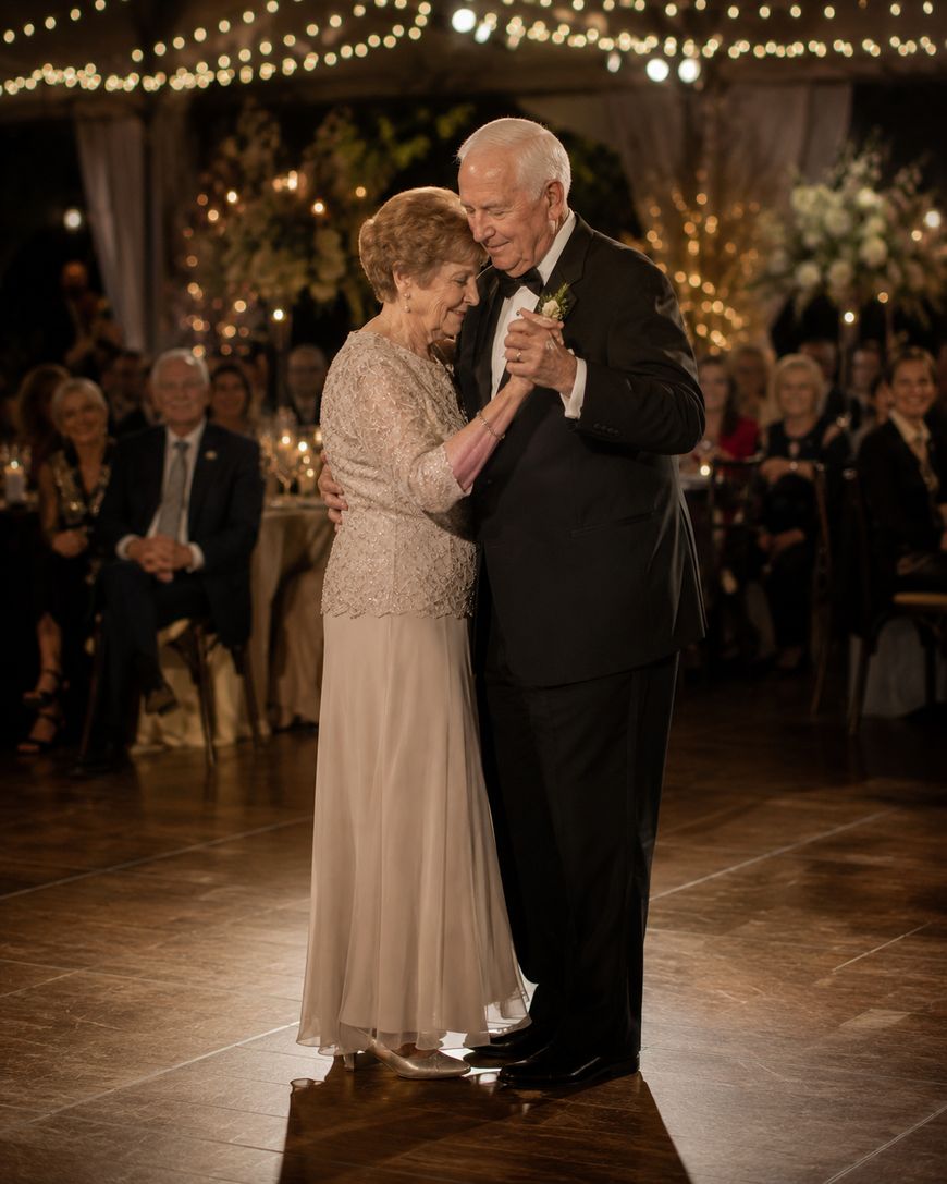 An elderly couple dressed elegantly shares a slow dance under string lights at a formal event, surrounded by seated guests who watch and smile warmly.