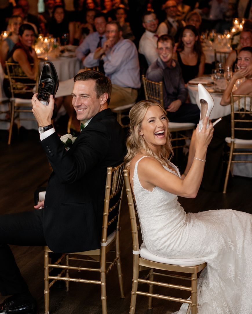 A bride and groom sit back-to-back on chairs at their wedding reception, each holding up one of the others shoes, smiling and laughing as guests seated at round tables watch and enjoy the moment.