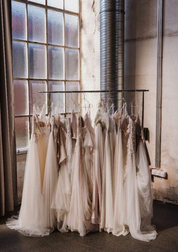 A clothing rack stands by a large industrial window, displaying several white and blush pink tulle dresses, including wedding gowns, in a softly lit room with exposed pipes and concrete walls.