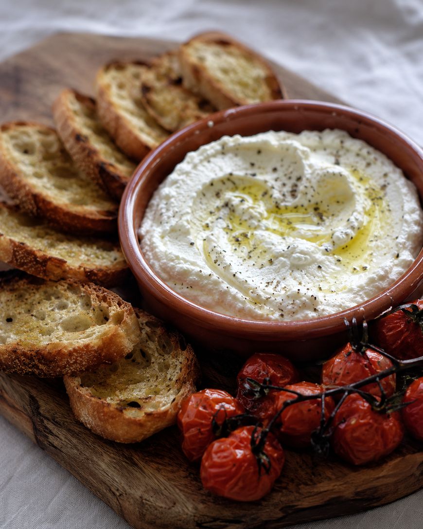 A wooden board with crispy toasted bread slices, a bowl of creamy whipped ricotta drizzled with olive oil and cracked black pepper, and roasted vine tomatoes.