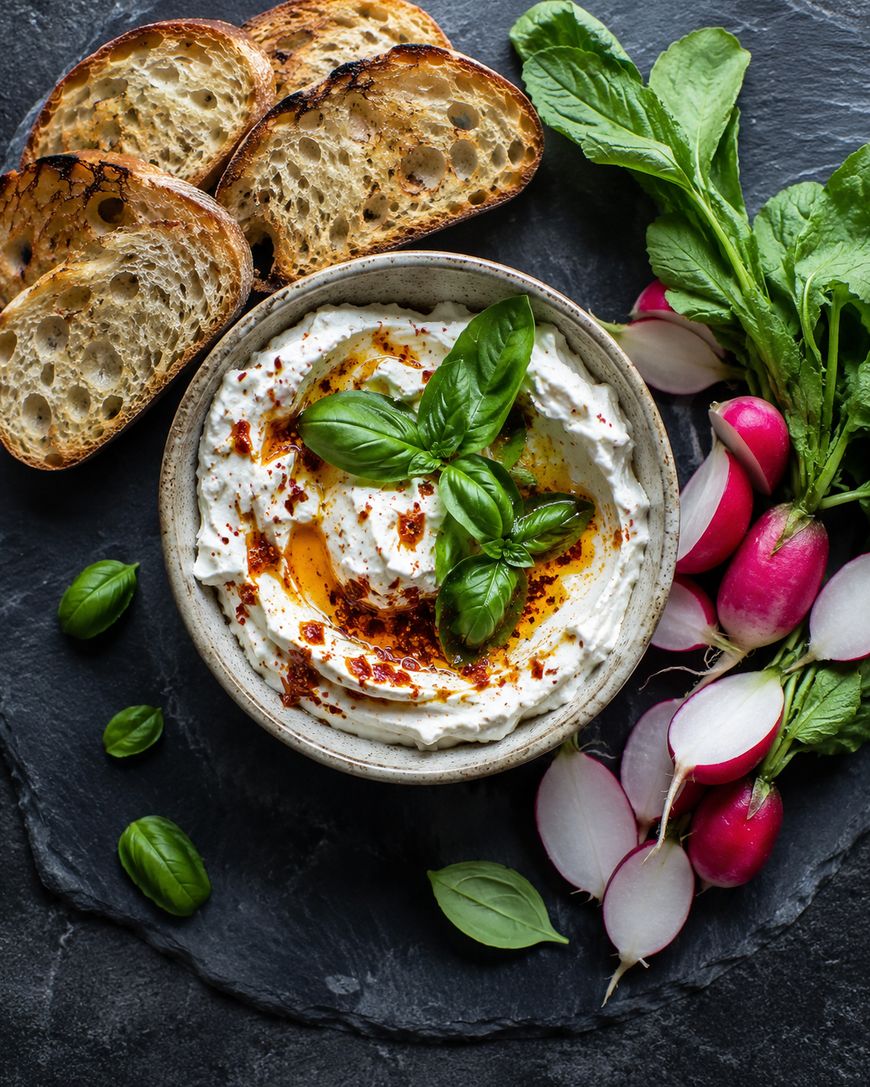 A bowl of creamy dip garnished with basil, olive oil, and spices, surrounded by slices of toasted bread, fresh radishes, and basil leaves on a dark slate surface.