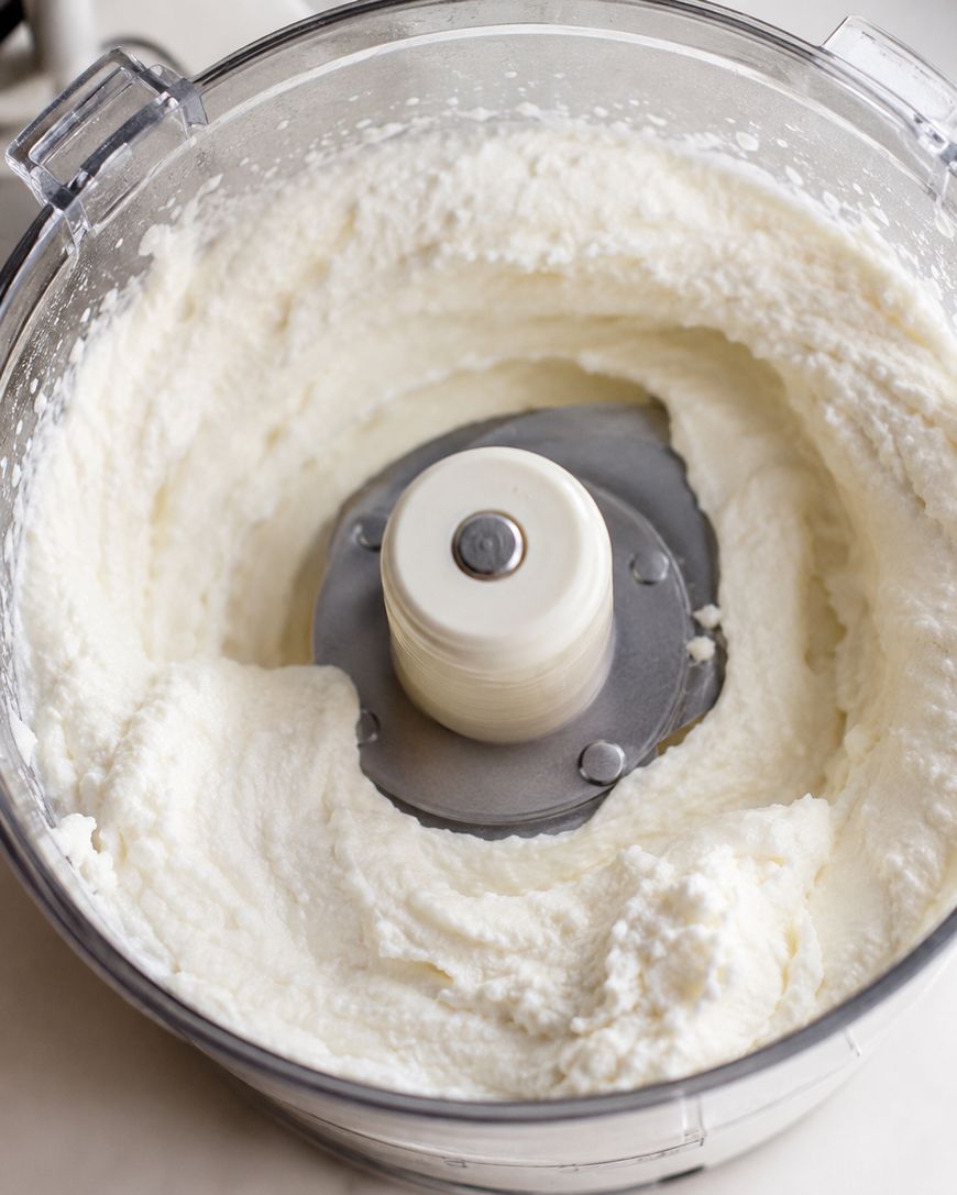 A food processor bowl filled with smooth, creamy white ricotta cheese mixture, with the blade visible in the center.