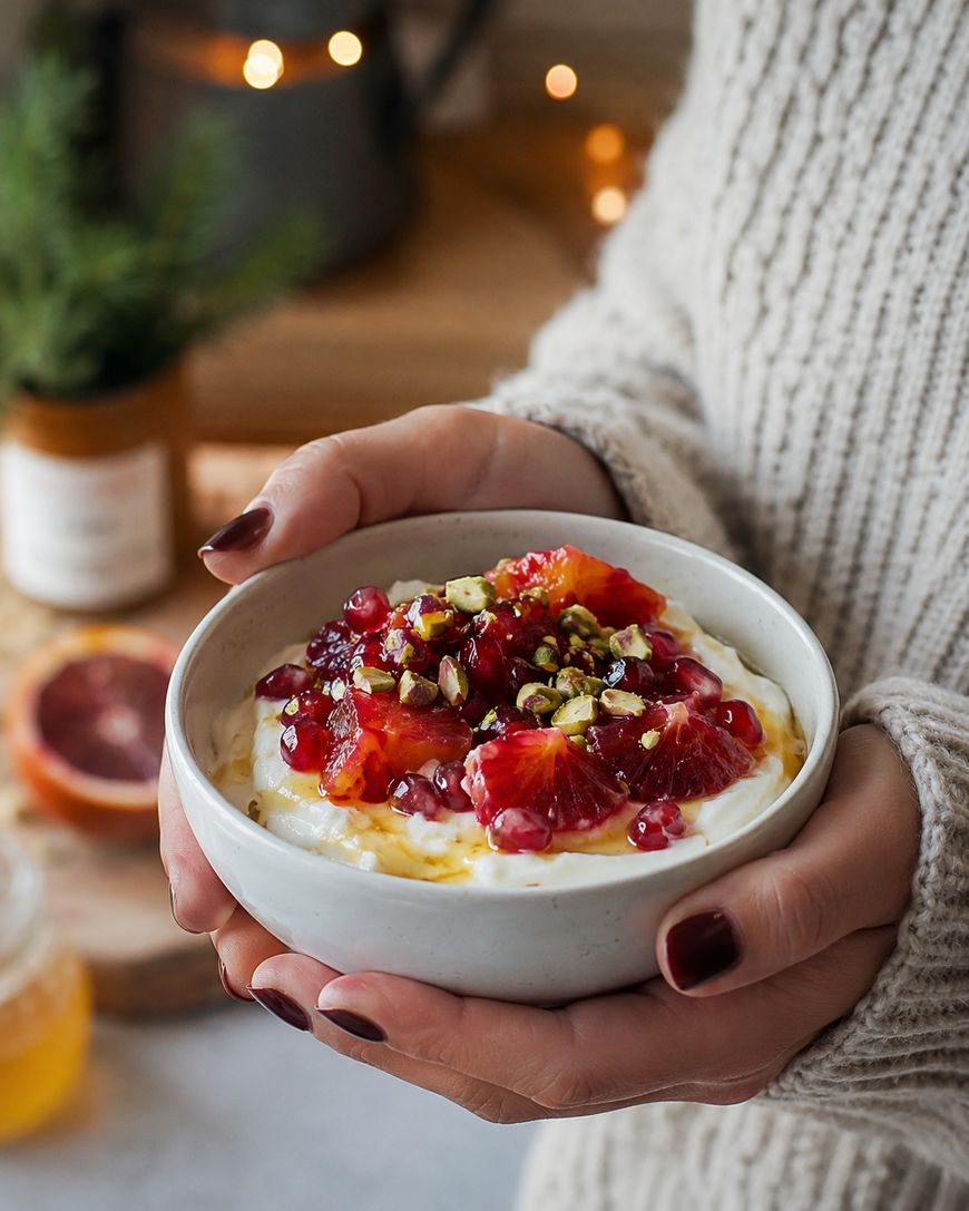 A person in a cozy sweater holds a bowl of yogurt topped with blood orange slices, pomegranate seeds, chopped pistachios, and a drizzle of honey. Warm, softly lit kitchen background with blurred decor.