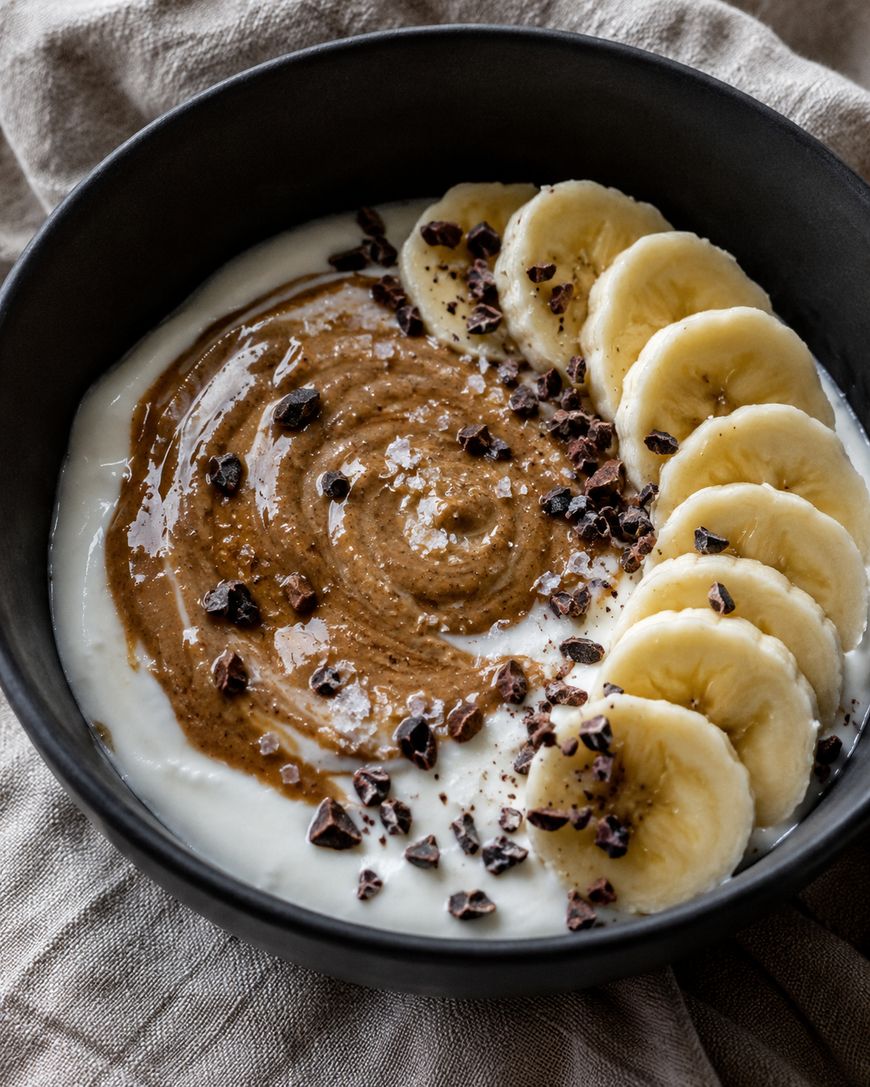 A black bowl filled with yogurt, swirled with nut butter, topped with a row of banana slices and sprinkled with cacao nibs, set on a beige cloth background.
