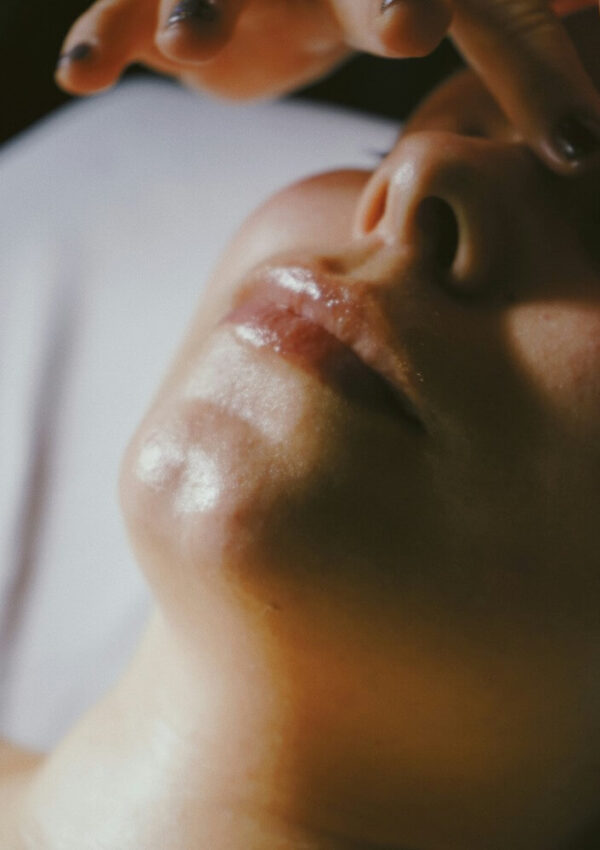 A close-up of a person’s face lying down, with a shiny, moisturized chin and lips. Fingers with dark nail polish are near their forehead, possibly during a skincare treatment or facial.