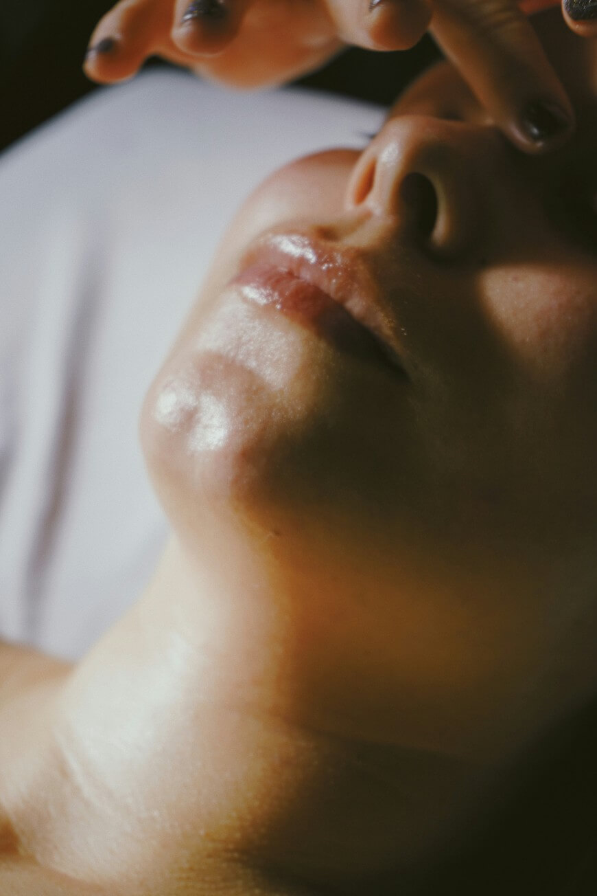 A close-up of a persons face and chin with glossy skin, as a hand with painted nails gently touches their forehead. The lighting is soft and warm.