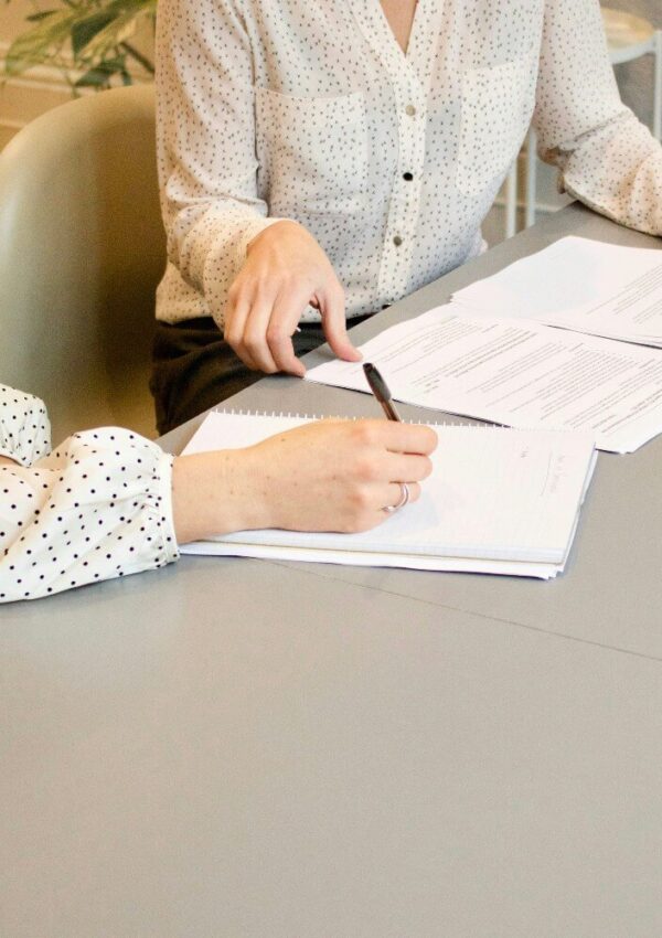 Two people sitting at a table, reviewing papers and taking notes. Both are wearing white, polka-dotted shirts. Only their hands and torsos are visible. The table is gray and there are plants in the background.