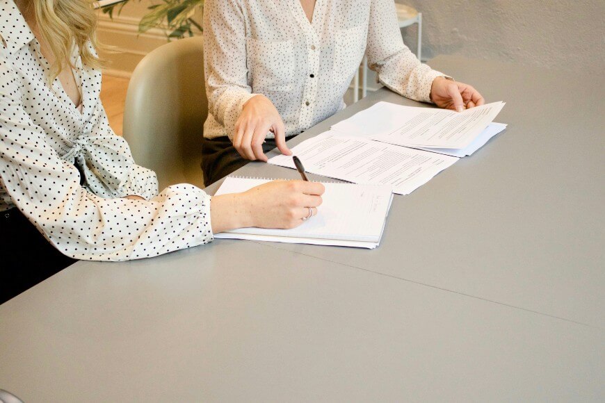 Two women in polka dot blouses sit at a table, reviewing and writing on paperwork together. Only their torsos and arms are visible.