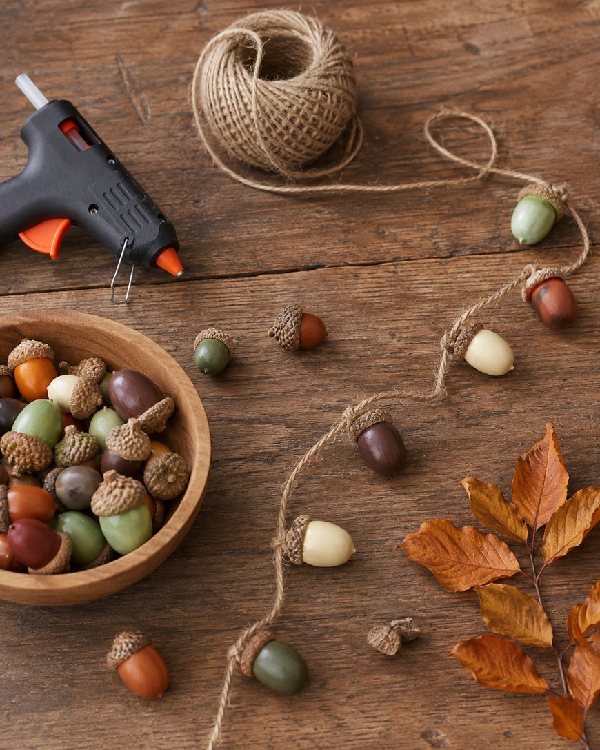 A rustic table with a glue gun, twine, a bowl of acorns, artificial acorns strung on twine, and brown leaves, suggesting a fall craft project in progress.