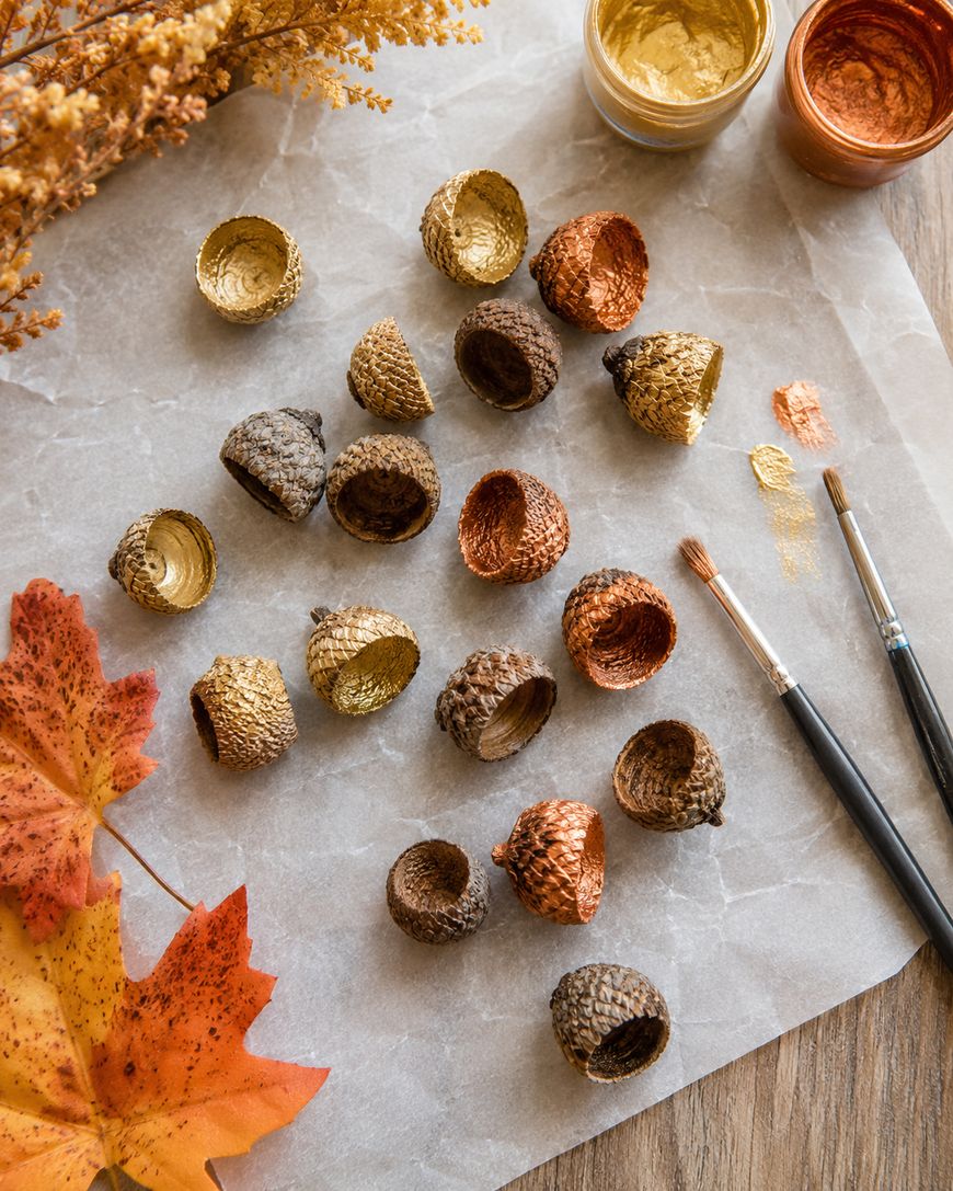 Acorn caps painted in gold, bronze, and natural colors are scattered on parchment paper. Two paintbrushes, paint jars, autumn leaves, and dried flowers are arranged around them, creating a fall craft scene.