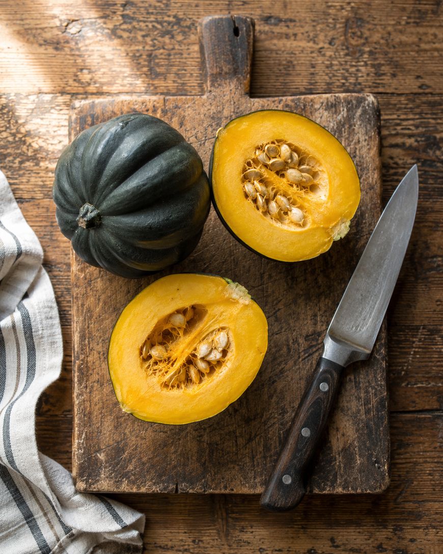 A whole acorn squash and one cut in half sit on a wooden cutting board next to a knife; a striped cloth is nearby, all on a rustic wooden table.