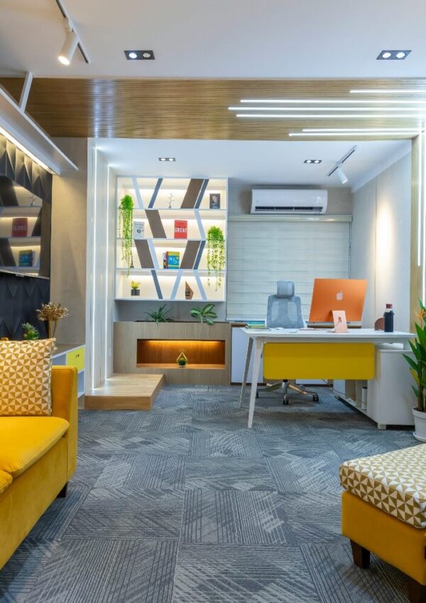 Modern office with a yellow sofa, geometric bookshelf, desk with an iMac, potted plants, gray carpet, and stylish lighting.