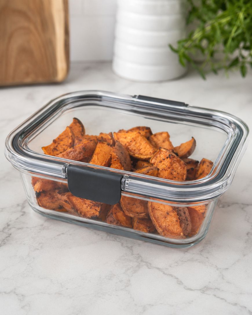 Glass container filled with roasted sweet potato wedges sits on a marble countertop, with a wooden cutting board and green herbs in the background.