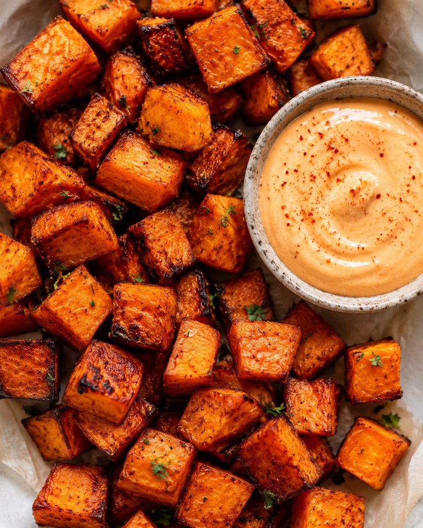 Roasted sweet potato cubes garnished with herbs, served on parchment paper next to a small bowl of creamy dipping sauce sprinkled with paprika.