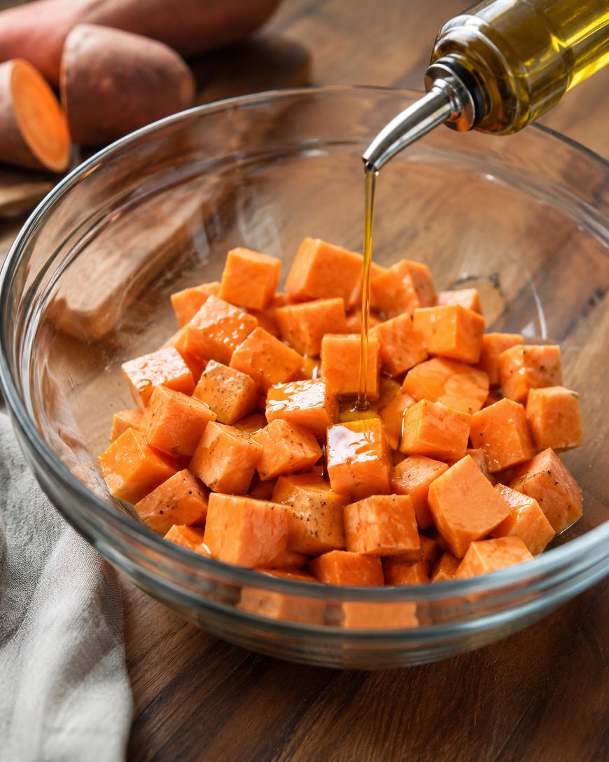 Olive oil being poured onto cubed sweet potatoes in a clear glass bowl, with whole sweet potatoes and a kitchen towel on a wooden surface in the background.