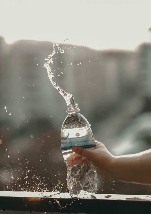 A hand squeezes a plastic water bottle, causing water to spray upward in a twisting splash against a blurred outdoor background.