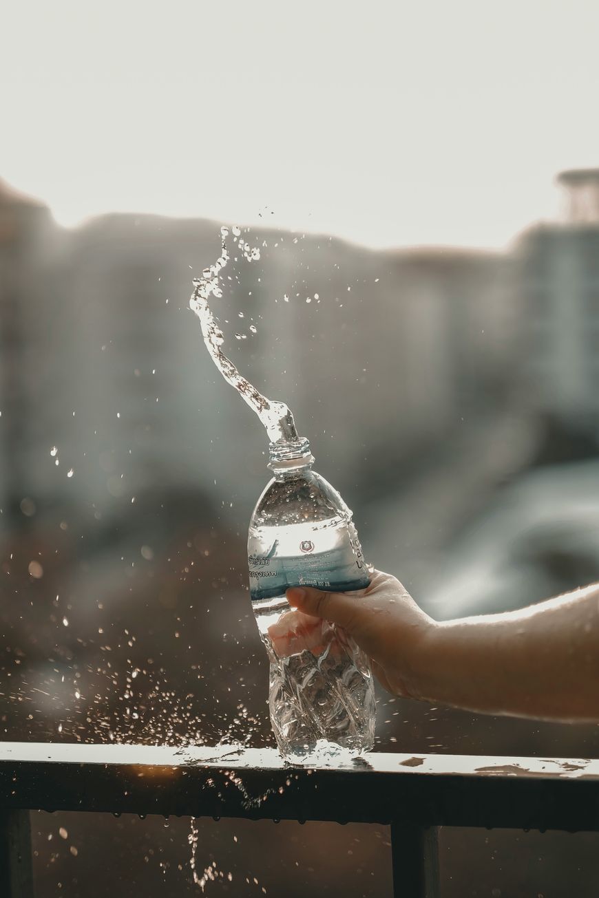 A hand squeezes a plastic water bottle, causing water to splash upward in a curved shape, with a blurred urban background and railings below.
