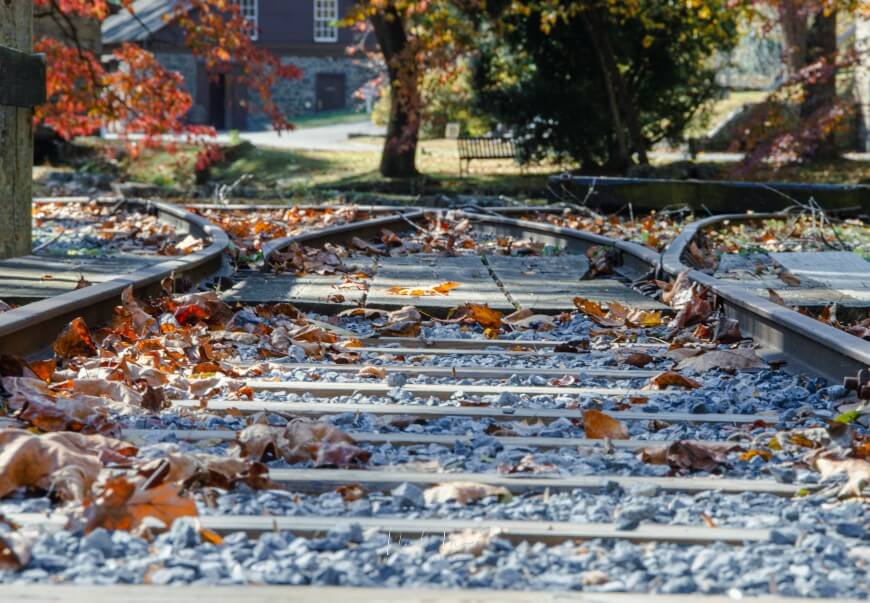 Close-up view of railway tracks covered with fallen autumn leaves, with trees and a building in the blurred background. The tracks split into two directions, and sunlight filters through the trees.