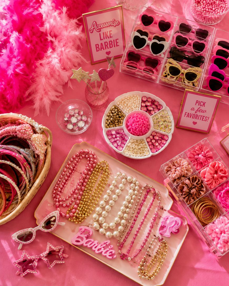 A pink-themed Barbie accessories display with beaded necklaces, sunglasses, headbands, hair ties, star wands, and signs reading “Accessorize like Barbie” and “Pick a few favorites.” Everything is arranged on a pink table.