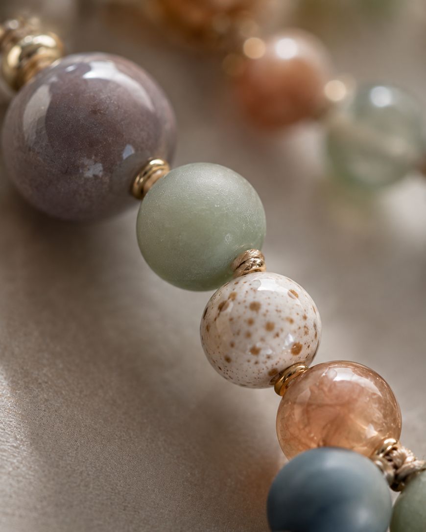 Close-up of a beaded necklace with round beads in various colors and textures, including green, speckled white, brown, and translucent amber, separated by small gold spacers on a soft beige background.