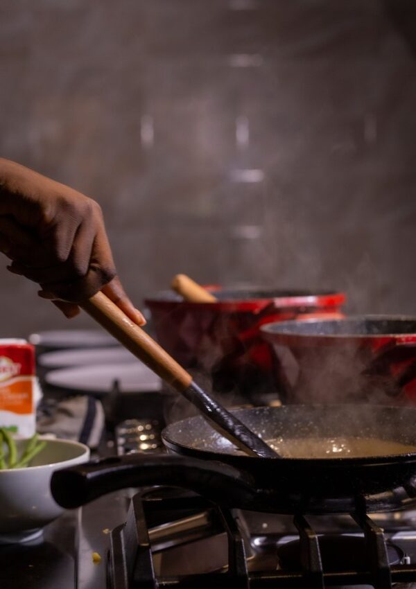 A hand stirs food in a steaming pan on a stovetop, with red pots and a bowl of green vegetables nearby.