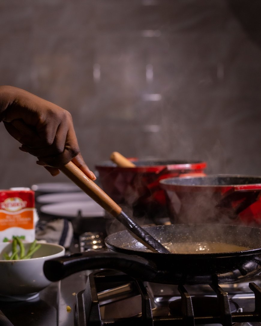 A hand stirs food in a frying pan on a stove, with steam rising. Red pots, a bowl with green beans, and a carton are nearby in a dimly lit kitchen.