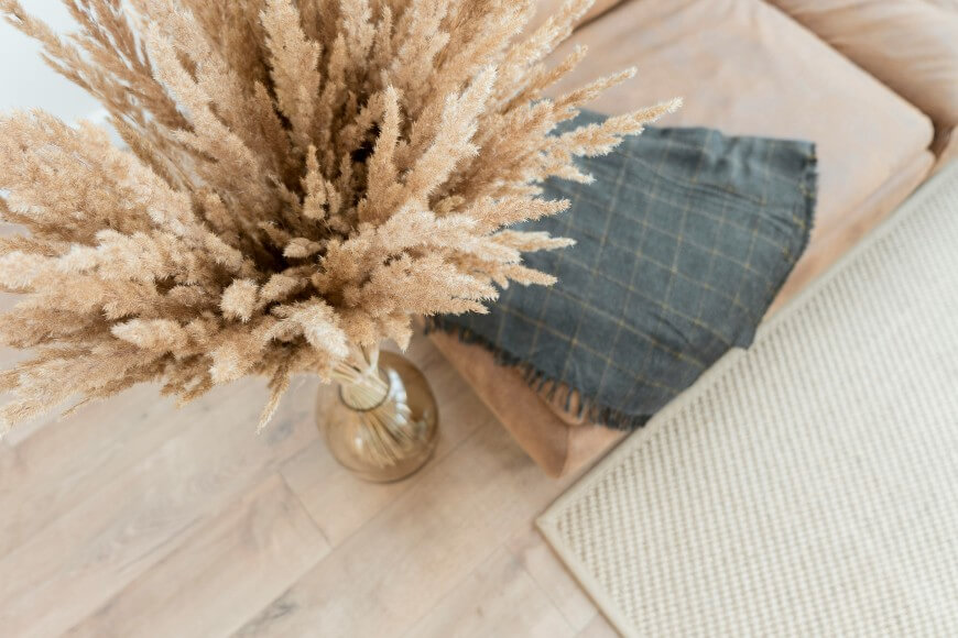 A vase with dried pampas grass sits on a light wooden floor next to a beige sofa with a gray plaid throw and a textured cream rug.