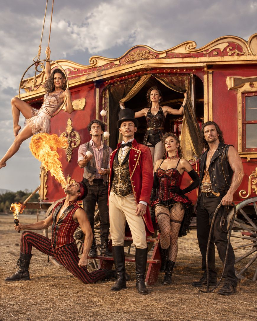 A circus troupe poses in front of a red wagon; a fire breather blows flames, an aerialist hangs from a hoop, and others wear theatrical costumes, including a ringmaster in a red coat, all standing on dry ground under a cloudy sky.