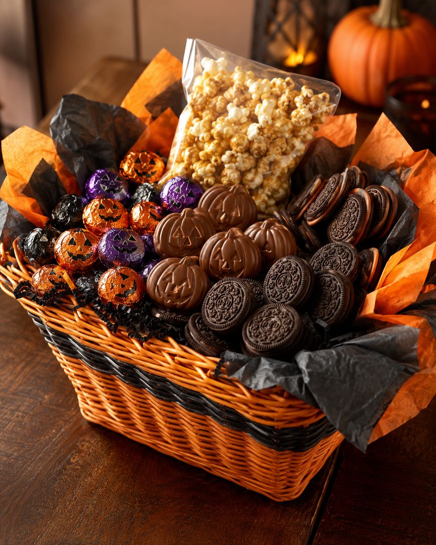A wicker basket filled with caramel popcorn, chocolate cookies, chocolate pumpkins, and orange and purple foil-wrapped chocolates, decorated with orange and black tissue paper.