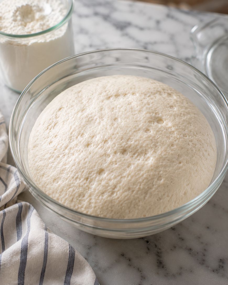 A glass bowl filled with risen bread dough sits on a marble countertop next to a jar of flour, a kitchen towel, and a glass lid.