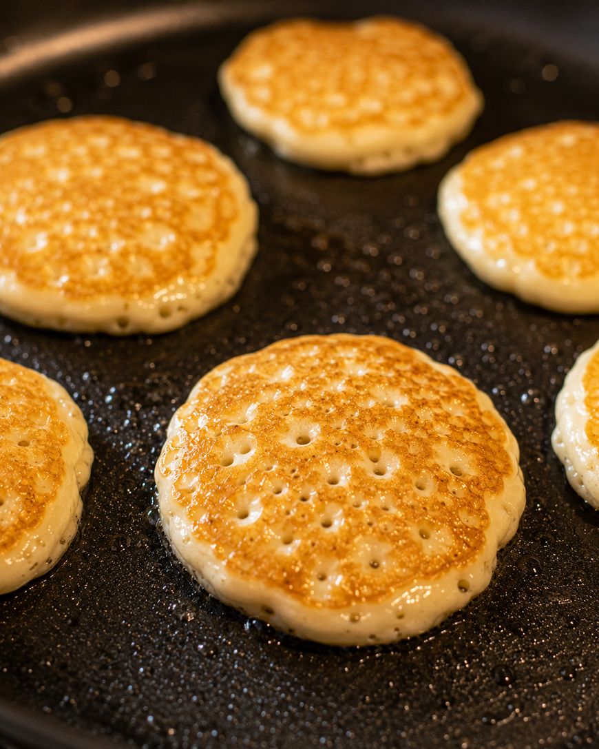 Six golden pancakes are cooking on a greased skillet, with bubbles visible on their surfaces, indicating they are freshly made and mid-cooking.