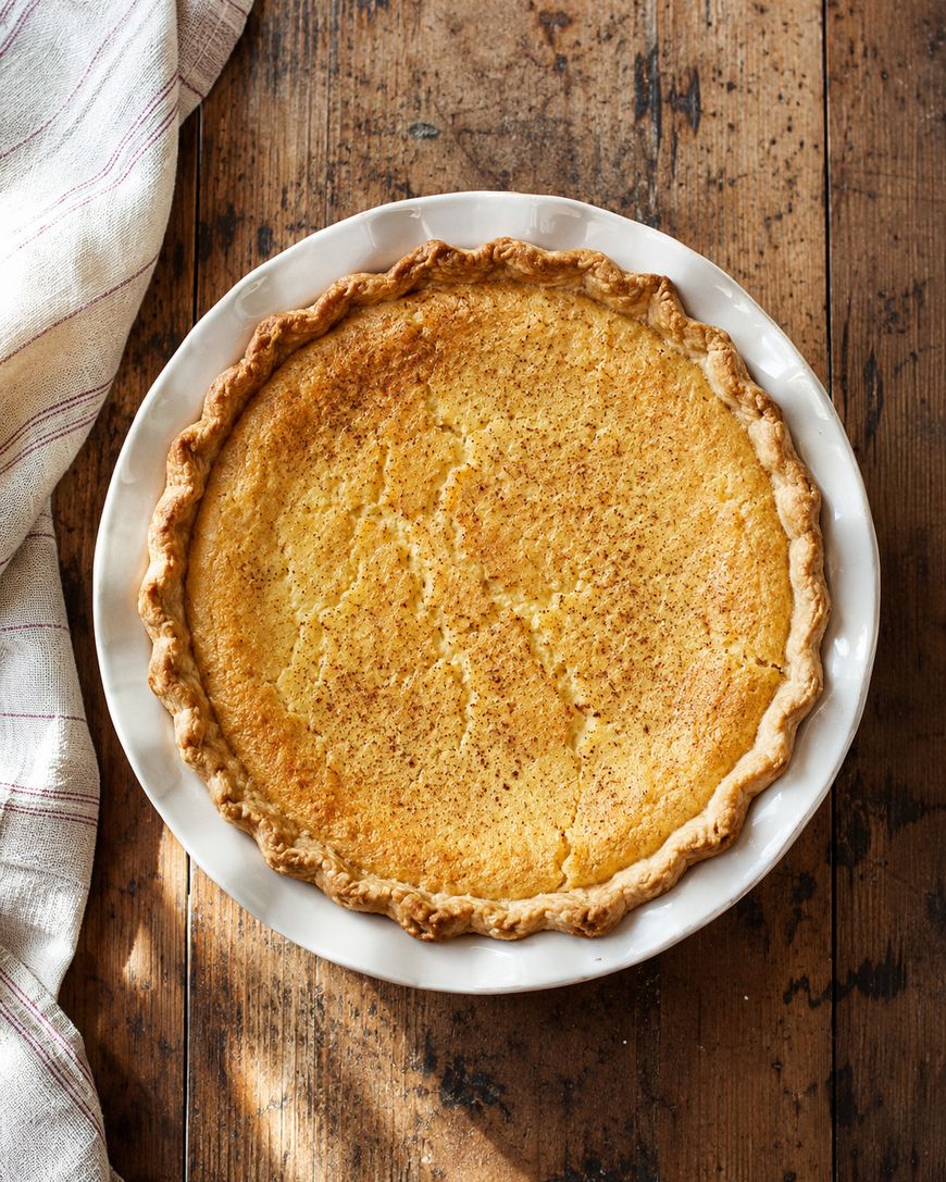A golden-brown chess pie with a flaky crust sits in a white pie dish on a rustic wooden table, next to a folded striped cloth.