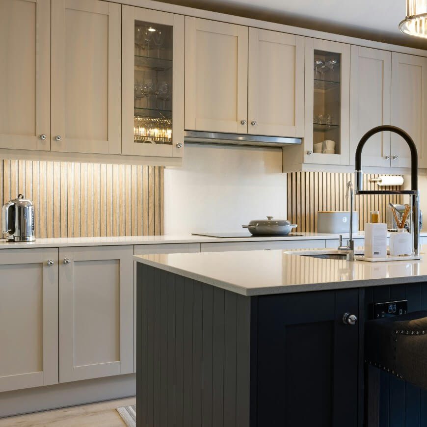 Modern kitchen with cream cabinets, glass doors, a dark island, built-in stove, and stainless steel faucet. Under-cabinet lighting highlights a vertical wood backsplash. Countertops are light and uncluttered.