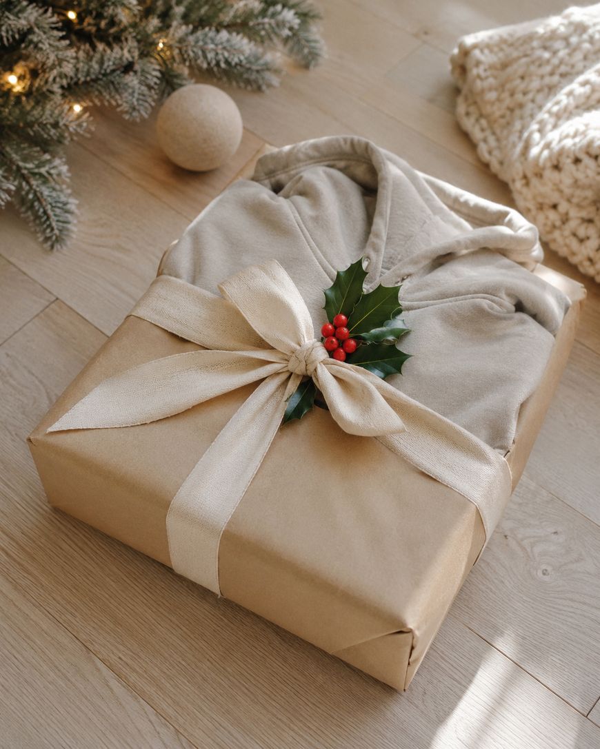 A gift wrapped in brown paper with a cream ribbon and a holly sprig. A beige hoodie is partially visible inside. The present sits on a wooden floor near a Christmas tree and a knitted blanket.