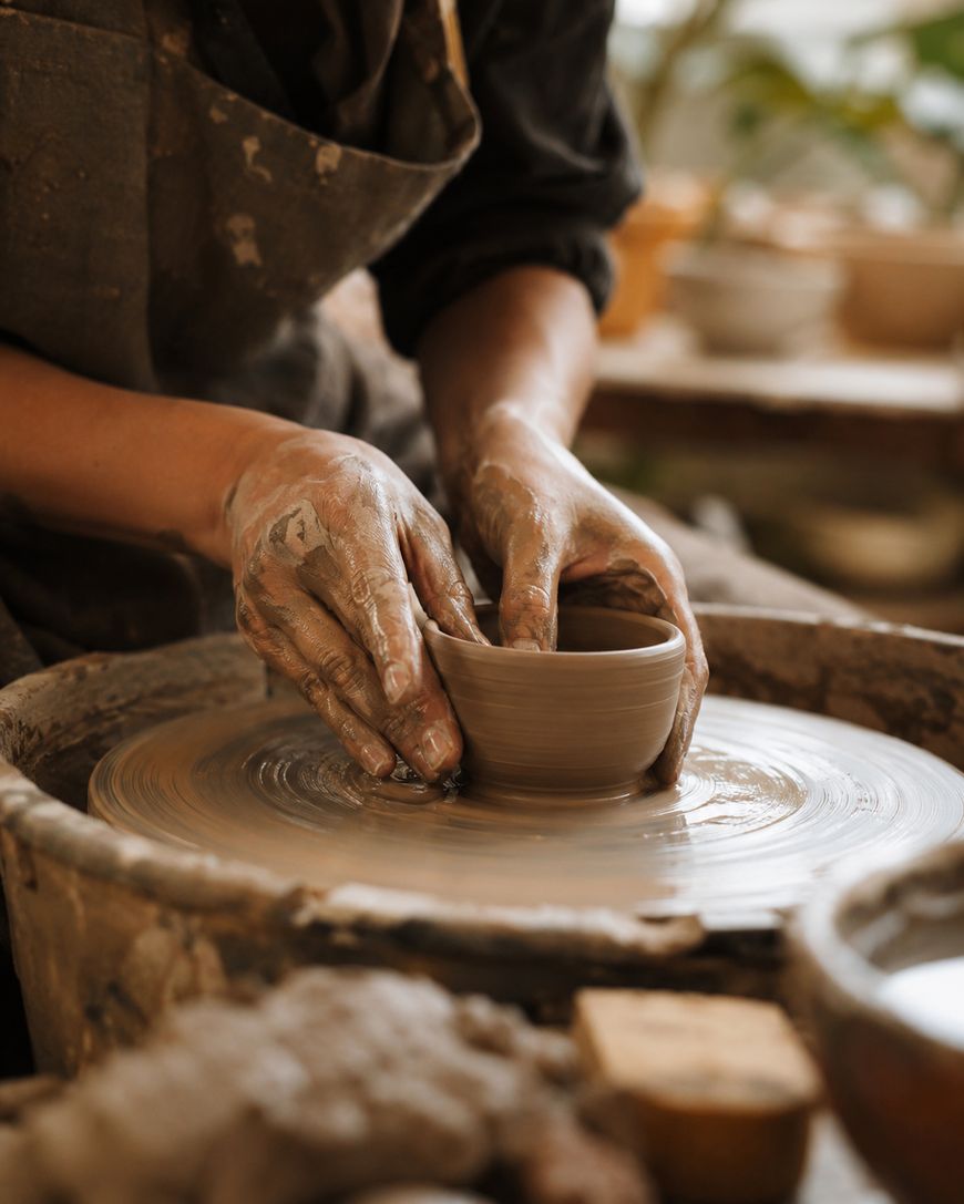 A person shapes a small clay bowl on a spinning pottery wheel, their hands covered in wet clay, in a warm, sunlit studio.